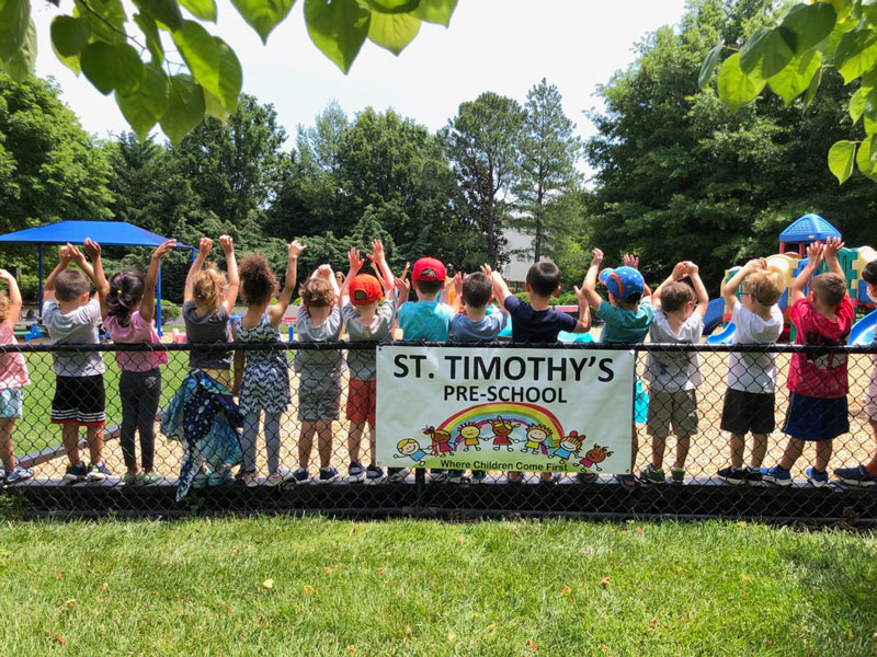 Kids standing along a fence with a St Timothy's Preschool Banner